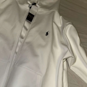 Vit hoodie från Ralph Lauren - Säljer en stilren vit hoodie från Ralph Lauren med dragkedja och det klassiska logotypbroderiet på bröstet. Perfekt för en avslappnad och snygg look. Hör av dig vid intresse! Storlek M-L