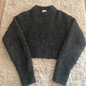 Mörkgrå croppad stickad tröja - Säljer en mysig mörkgrå croppad stickad tröja med långa ärmar och ribbade detaljer vid ärmslut och nederkant. Perfekt för kyliga dagar!