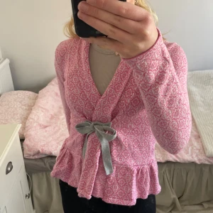 Rosa mönstrad kofta från Odd Molly - Säljer en söt rosa kofta från Odd Molly med ett unikt mönster och broderad detalj på ryggen. Koftan har långa ärmar och en knytning framtill med volangdetalj vid nederkanten. Perfekt för att lägga till en färgglad touch till din outfit!