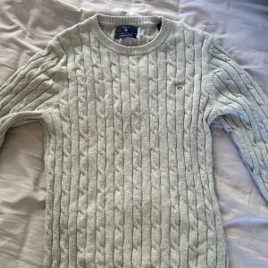 Grå kabelstickad tröja från GANT - Säljer en stilren grå kabelstickad tröja från GANT i stretchig bomull. Tröjan har en rund halsringning och ett diskret broderat emblem på bröstet. Perfekt för en klassisk och bekväm look.