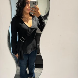 Svart omlottopp med bälte - Snygg svart omlottopp med bälte och v-ringning. Toppen har långa ärmar och en stilren design som passar perfekt till både jeans och kjol.