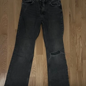 Svarta jeans med slitningar - Snygga svarta jeans med en cool slitning på ena benet. De har en bootcut-stil som ger en retro känsla. Perfekta för en avslappnad look.