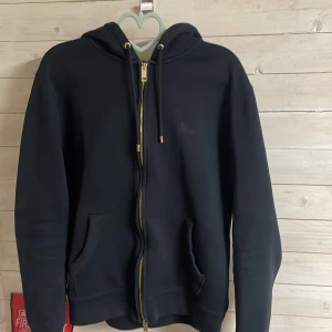 Svart hoodie från Burberry - Säljer en stilren svart hoodie från Burberry med dragkedja och huva. Insidan av huvan har ett klassiskt Burberry-mönster i rött och grått. Tillverkad i bomull och polyester för en bekväm passform. Perfekt för en avslappnad look. L men passar liten M till stor S typ.