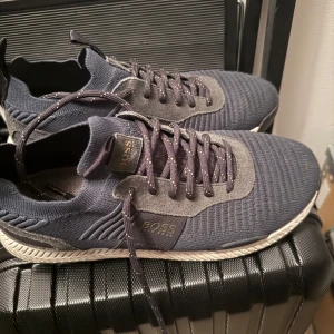 Mörkblå sneakers från Boss - Snygga mörkblå sneakers från Boss med stickad ovandel och grå detaljer. Skorna har snörning och en bekväm sula som ger bra stöd. Perfekta för en stilren och avslappnad look.