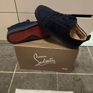 Christian Louboutin marinblå - Säljer ett par stilrena svarta sneakers från Christian Louboutin med den ikoniska röda sulan. Skorna har snörning och är tillverkade i ett elegant material som ger en lyxig känsla. Perfekta för dig som vill ha en touch av exklusivitet i vardagen.