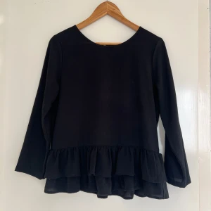 Svart topp med volang från ONLY - Säljer en stilren svart topp från ONLY med volangdetalj i nederkant. Toppen har långa ärmar och en dragkedja i ryggen. Perfekt som festtopp🖤