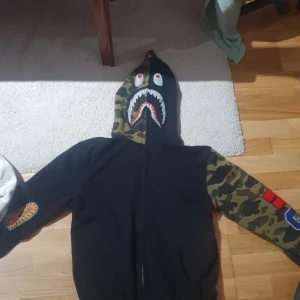 Svart camo hoodie från A Bathing Ape - Säljer en cool svart hoodie från A Bathing Ape med kamouflagemönstrade ärmar och huva. Hoodien har en dragkedja framtill och ett hajmotiv på huvan. Perfekt för streetwear-entusiaster!