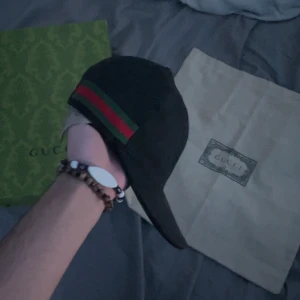 Svart keps från Gucci - Snygg svart keps från Gucci med klassiska gröna och röda ränder på sidan. Perfekt för att ge din outfit en lyxig touch. Kommer med originalförpackning och dustbag.