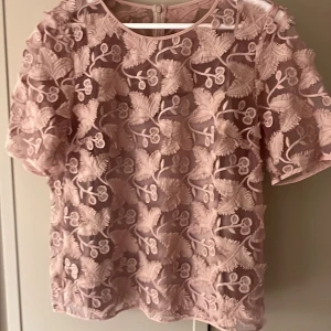 Rosa blus med blommigt mönster från Baun undpferdgarten  - Säljer en söt rosa blus med ett vackert blommigt mönster. Blusen har korta ärmar och en rund halsringning. Perfekt för en somrig look!