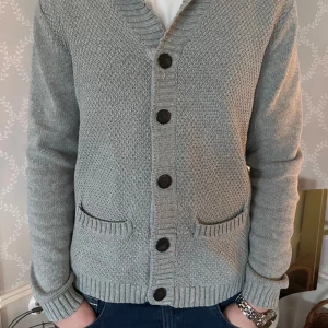 Grå stickad kofta med knappar - Säljer en stilren grå stickad kofta med knappar framtill. Koftan har långa ärmar och ribbade muddar vid ärmslut och nederkant. Perfekt för kyligare dagar. Koftan är från märket mango. Modellen är runt 180 och väger 75kg. 
