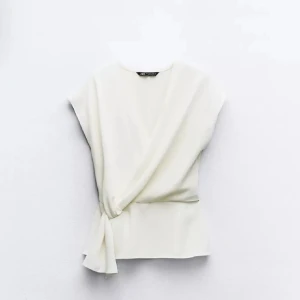 Vit omlottopp från Zara - Elegant vit omlottopp från Zara med knytning i sidan. Perfekt för en stilren look. Toppen har en lös passform och är ärmlös, vilket gör den idealisk för varmare dagar.