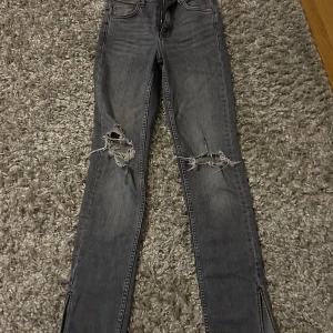 Grå jeans med slitningar från Zara - Snygga grå jeans från Zara med coola slitningar på knäna. De har en klassisk femficksdesign och knappgylf. Perfekta för en avslappnad stil med en edgy touch.