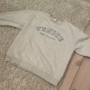 Grå sweatshirt med tryck - Säljer en grå sweatshirt med texten 'VENICE Caffè Panorama' i blått på framsidan. Tröjan har långa ärmar och ribbade muddar vid ärmslut och nederkant. Använd typ 5 gånger.Perfekt för en avslappnad stil.