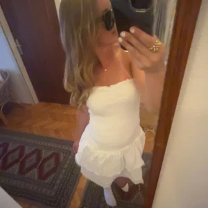 Vit axelbandslös klänning från Gina - Säljer en söt vit axelbandslös klänning med volangdetaljer. Perfekt för sommarens alla tillfällen. Köpte den i L men passar perfekt till den som har storlek M.