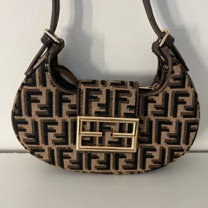 Beige och svart axelväska med monogram - Snygg axelväska i beige och svart. Väskan har en praktisk dragkedja och ett elegant spänne framtill. Perfekt för att ge en lyxig touch till din outfit.