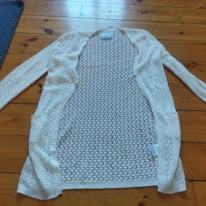 Vit stickad kofta från Vintage Skill - Säljer en vit stickad kofta från Vintage Skill. Den har ett fint hålmönster och långa ärmar. Perfekt för lager-på-lager-stil. Koftan har två fickor framtill och ribbade kanter.