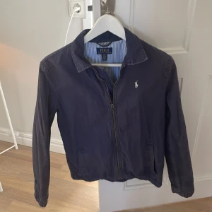 Blå jacka från Polo Ralph Lauren - Säljer en stilren blå jacka från Polo Ralph Lauren med dragkedja och broderad logga på bröstet. Jackan har en klassisk krage och långa ärmar. Perfekt för en avslappnad och snygg look.
