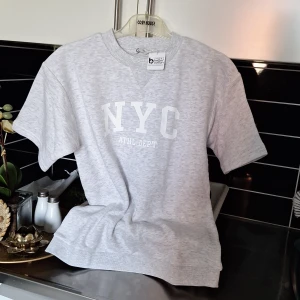 Grå t-shirt med tryck från Cubus - Säljer en stilren grå t-shirt från Cubus med texten 'NYC ATHL. DEPT' tryckt i vitt på framsidan. T-shirten är kortärmad och tillverkad i bomull, perfekt för en avslappnad look.