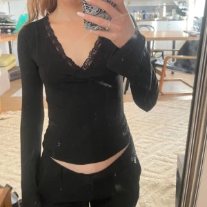 Svart långärmad topp med spetsdetaljer - Populär tröja från Nelly. Snygg svart långärmad topp med spetsdetaljer vid halsringningen. Perfekt för en stilren look. Toppen har en v-ringad design som ger en elegant touch. Använd en gång. 