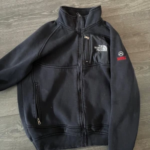 Svart fleecejacka från The North Face - Säljer en svart fleecejacka från The North Face med dragkedja och broderad logga på bröstet. Jackan har långa ärmar och ribbade muddar. Perfekt för kyliga dagar.