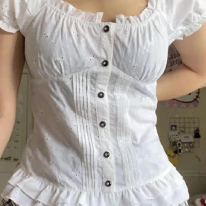 Vit blus med knappar och snörning - Charmig vit blus med korta ärmar och dekorativa knappar framtill. Blusen har ett broderat mönster och snörning i ryggen för en justerbar passform. 