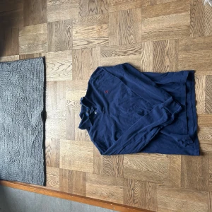 Ralph Lauren Blå pikétröja  - Säljer en klassisk blå pikétröja från Ralph Lauren med röd broderad logga på bröstet. Tröjan har långa ärmar och en stilren krage med knappar. Perfekt för en avslappnad men ändå stilfull look.