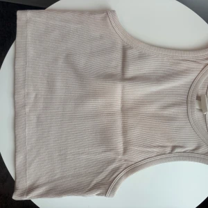 Beige ribbad linnetopp från H&M - Säljer en stilren beige ribbad linnetopp från H&M. Perfekt för varma dagar med sin ärmlösa design och mjuka material. Enkel att matcha med olika outfits. Passar perfekt för en avslappnad look.