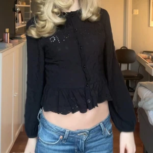 Svart blus med spetsdetaljer - Snygg svart blus med spetsdetaljer och knappar framtill. Blusen har långa ärmar och en volangkant nedtill som ger en feminin touch. Perfekt för att klä upp en vardagslook eller för en kväll ute.