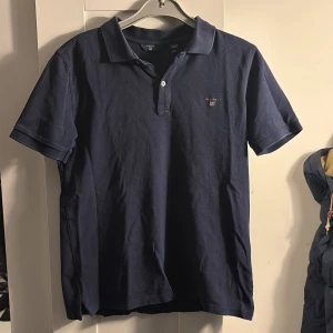 Gant pikétröja  - Säljer en stilren mörkblå pikétröja från GANT. Tröjan har en klassisk krage med två knappar och det ikoniska GANT-emblemet broderat på bröstet. Storlek 158/164cm, 13-14 år antar att det kan vara storlek xs och s