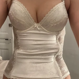 Vit korsett med spetsdetaljer - Säljer en vit korsett med spetsdetaljer och justerbara axelband. Den har en vacker broderad design och man kan knäppa den på tre olika rader beroende på hur man vill att den ska sitta. Perfekt för att ge en lyxig känsla till din outfit. Den korvar sig lite därfram