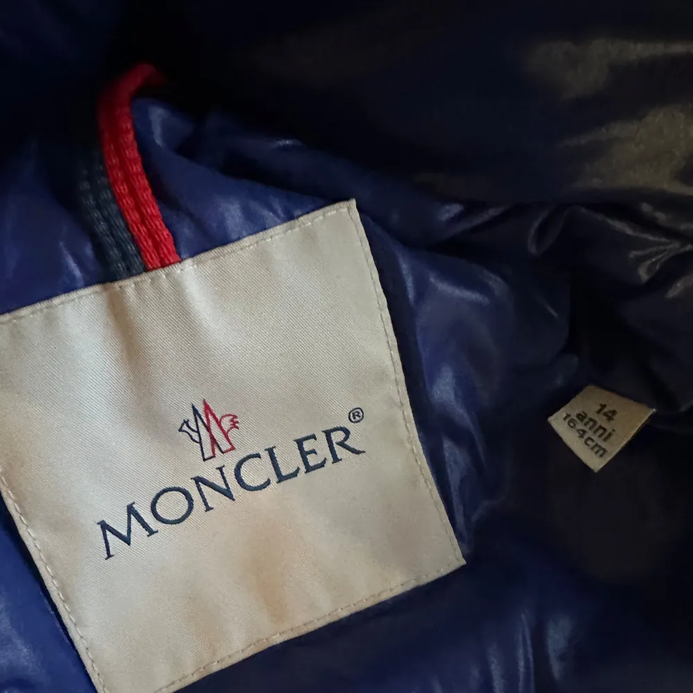 Säljer en snygg blå dunjacka från Moncler med huva och dragkedja. Jackan har långa ärmar och två fickor med dragkedja framtill. Perfekt för kyliga dagar.stl14/164. Takit.