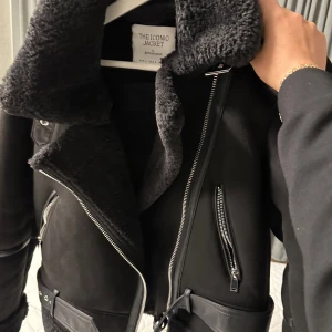 Svart skinnjacka med foder - Snygg svart skinnjacka med mjukt foder och flera dragkedjor. Jackan har en cool design med bälte i midjan och en rejäl krage. Perfekt för att hålla värmen och stilen under kyliga dagar.