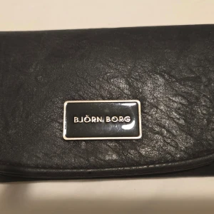 Svart plånbok från Björn Borg - Snygg svart plånbok från Björn Borg i skinn med flera fack för kort och en dragkedja för mynt. Perfekt för att hålla ordning på dina tillhörigheter. Stängs med tryckknapp och har en stilren design.