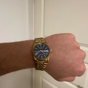 Guldfärgad klocka från Kompass - Snygg guldfärgad klocka från Kompass med blå urtavla och guldfärgade visare. Armbandet är i metall med en klassisk länkdesign. Perfekt för den som vill ha en stilren och elegant accessoar.