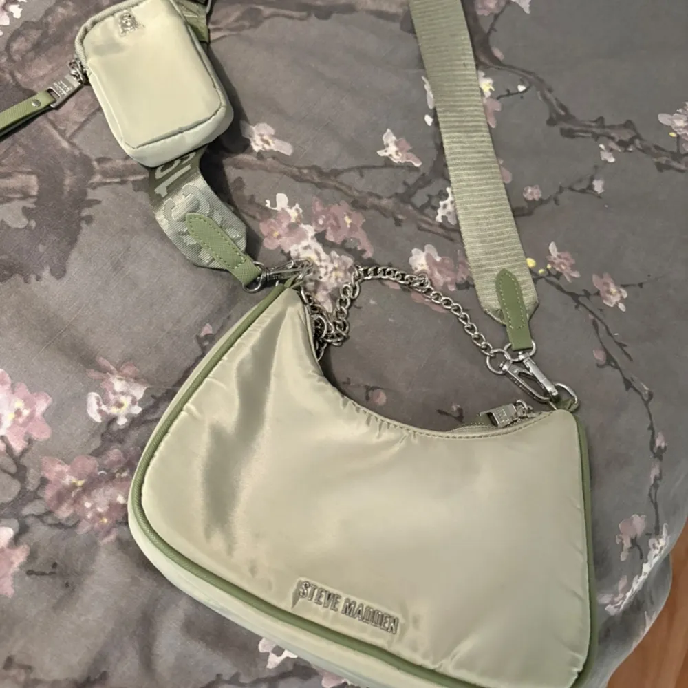 Snygg grön axelväska från Steve Madden med justerbar axelrem och kedjedetalj. Väskan har en praktisk liten ficka med dragkedja och en extra liten väska fäst vid remmen. . Laukut & Käsilaukut.