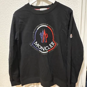 Svart tröja från Moncler - Säljer en svart tröja från Moncler med stort logotryck på framsidan. Tröjan har långa ärmar och en rund halsringning. Perfekt för en stilren och avslappnad look.