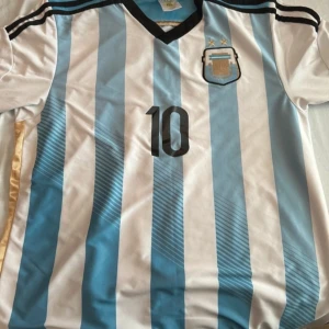 Argentina fotbollströja med nummer 10 - Säljer en klassisk Argentina fotbollströja från Adidas med nummer 10 på framsidan. Tröjan har ett randigt mönster i vitt och ljusblått med svarta detaljer vid kragen och axlarna. Perfekt för fotbollsfans!