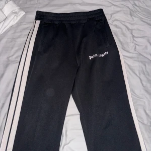Svarta byxor från Palm Angels - Snygga svarta byxor från Palm Angels med ljus beige ränder längs sidorna och logotypen tryckt på framsidan. Byxorna har en elastisk midja för extra komfort och är perfekta för en avslappnad stil. Kan användas överallt, antingen ha dom i skolan eller så har man dom hemma som ett par mjukis byxor, vid frågor skriv privat☺️