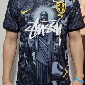 Svart Brasilien fotbollströja med grafiskt tryck - Snygg svart fotbollströja från Nike med Brasilien-tema. Tröjan har ett unikt grafiskt tryck med Kristusstatyn och texten 'Brasil' i gult. Kortärmad design med detaljer i blått och gult. Perfekt för fotbollsfans som vill sticka ut.