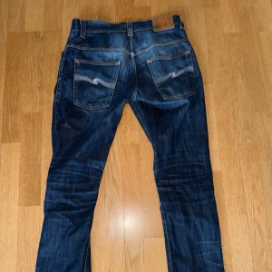 Nudie blå jeansbyxor med broderade bakfickor - Snygga blå jeans med broderade detaljer på bakfickorna från nudie. Klassisk femficksdesign med knapp och dragkedja framtill. Perfekta för en avslappnad stil. Byter och samfraktar