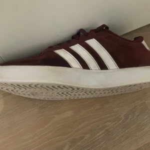 Vinröda sneakers från Adidas - Snygga vinröda sneakers från Adidas med klassiska vita ränder på sidan. Skorna har snörning och en vit sula som ger en stilren look. Perfekta för en avslappnad stil. Köpta på Adidas för 999kr