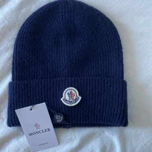 Mörkblå ribbad mössa från Moncler - Snygg mörkblå mössa från Moncler med ribbad design och uppvikt kant. Den har en broderad Moncler-logga framtill. Perfekt för att hålla värmen med stil.