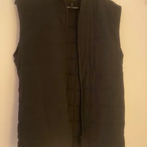 Svart quiltad väst från Rains - Snygg svart quiltad väst från Rains med dragkedja framtill. Perfekt för lager-på-lager under kyligare dagar. Enkel och stilren design som passar till det mesta.