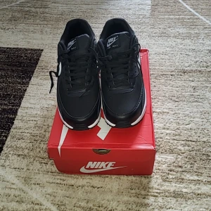 Svarta Nike Air Max - Snygga svarta Nike Air Max med vit sula och klassisk design. Skorna har snörning och den ikoniska Air Max-dämpningen för extra komfort. Perfekta för både stil och funktion.