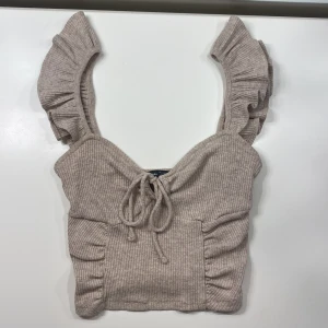Beige ribbad topp från Bershka - Säljer en beige ribbad topp från Bershka med volangdetaljer på axelbanden och knytning framtill. Perfekt för en stilren och trendig look🤗💕 köpte den i Polen för nått år sen men har inte används på länge 