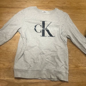 Grå sweatshirt från Calvin Klein - Säljer en stilren grå sweatshirt från Calvin Klein med deras ikoniska logga i svart på framsidan. Tröjan har långa ärmar och ribbade muddar vid ärmslut och nederkant. Perfekt för en avslappnad look.