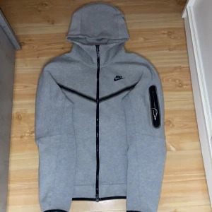 Grå huvtröja från Nike - Säljer en grå huvtröja från Nike med dragkedja och svarta detaljer. Tröjan har en stilren design med en huva och långa ärmar. Perfekt för en sportig look eller avslappnade dagar. 🖤den är äkta 