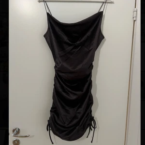Klänning  - Elegant svart/mörk brun klänning i siden med smala axelband och draperad design. Klänningen har en åtsittande passform och justerbara snören på sidorna. Som ny, använd 1-2 gånger. 
