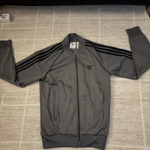 Grå träningszip från Adidas - Säljer en stilren grå träningszip från Adidas med de klassiska tre svarta ränderna längs ärmarna. Koftan har dragkedja framtill och en bekväm passform. Perfekt för träning eller en avslappnad stil. 