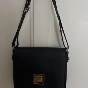 Svart axelväska med dragkedja - Snygg svart axelväska med justerbar rem och stilren design. Väskan har en praktisk dragkedja på baksidan och en fyrkantig metallspänne på framsidan. Perfekt för att bära dina dagliga nödvändigheter med stil.